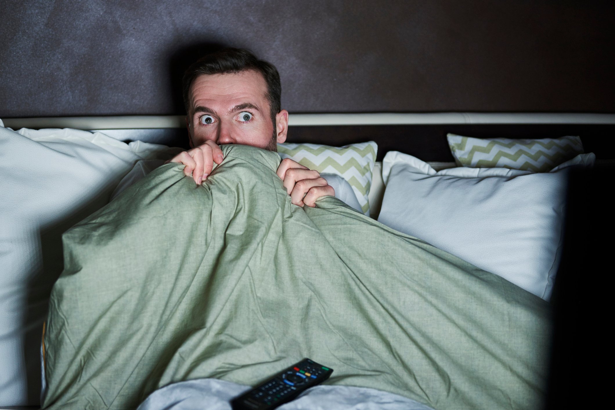 A wide-eyed man hides under a comforter on his bed while watching TV in a dark room.