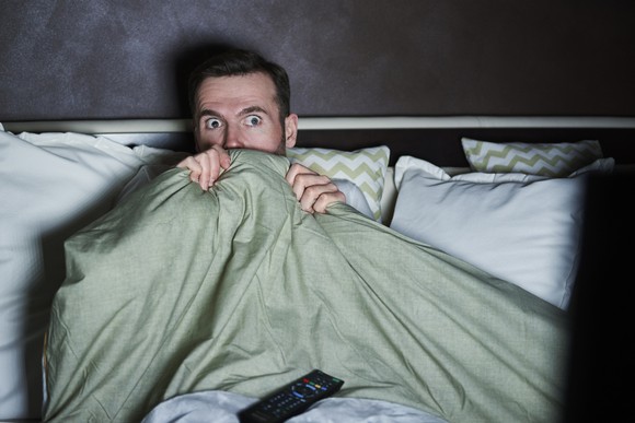 A wide-eyed man hides under a comforter on his bed while watching TV in a dark room.