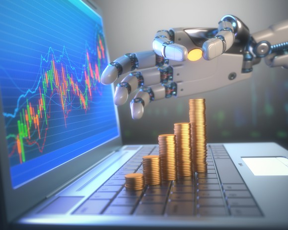A robotic hand adding a coin to a stack of coins on top of a laptop keyboard with a stock chart on the screen.