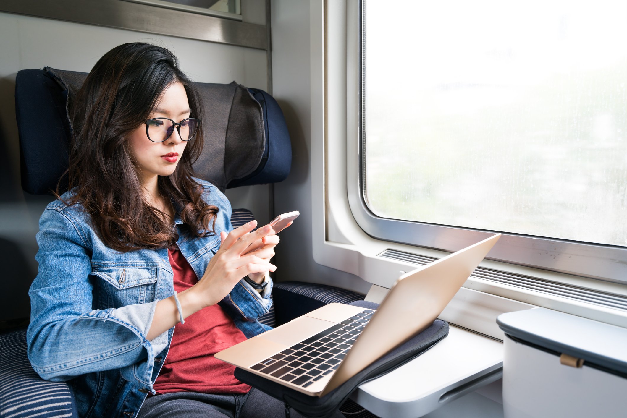 A young Asian woman on a train uses a laptop and a smartphone.