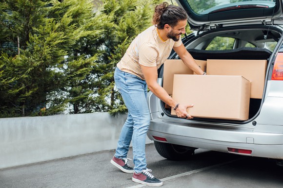 A young man in casual clothes loads large boxes into a hatchback car.
