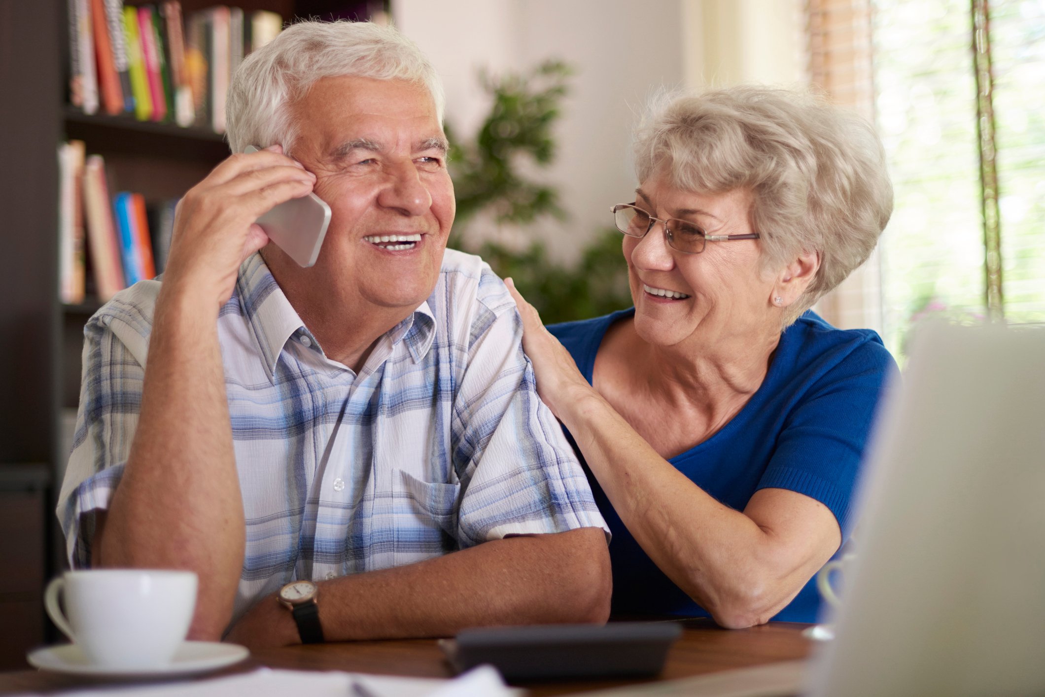 Older woman putting hand on older man's arm while he talks on the phone; both are smiling