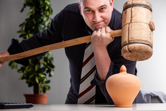 Smirking man in shirt and tie holding a large wooden mallet over a small pottery urn.