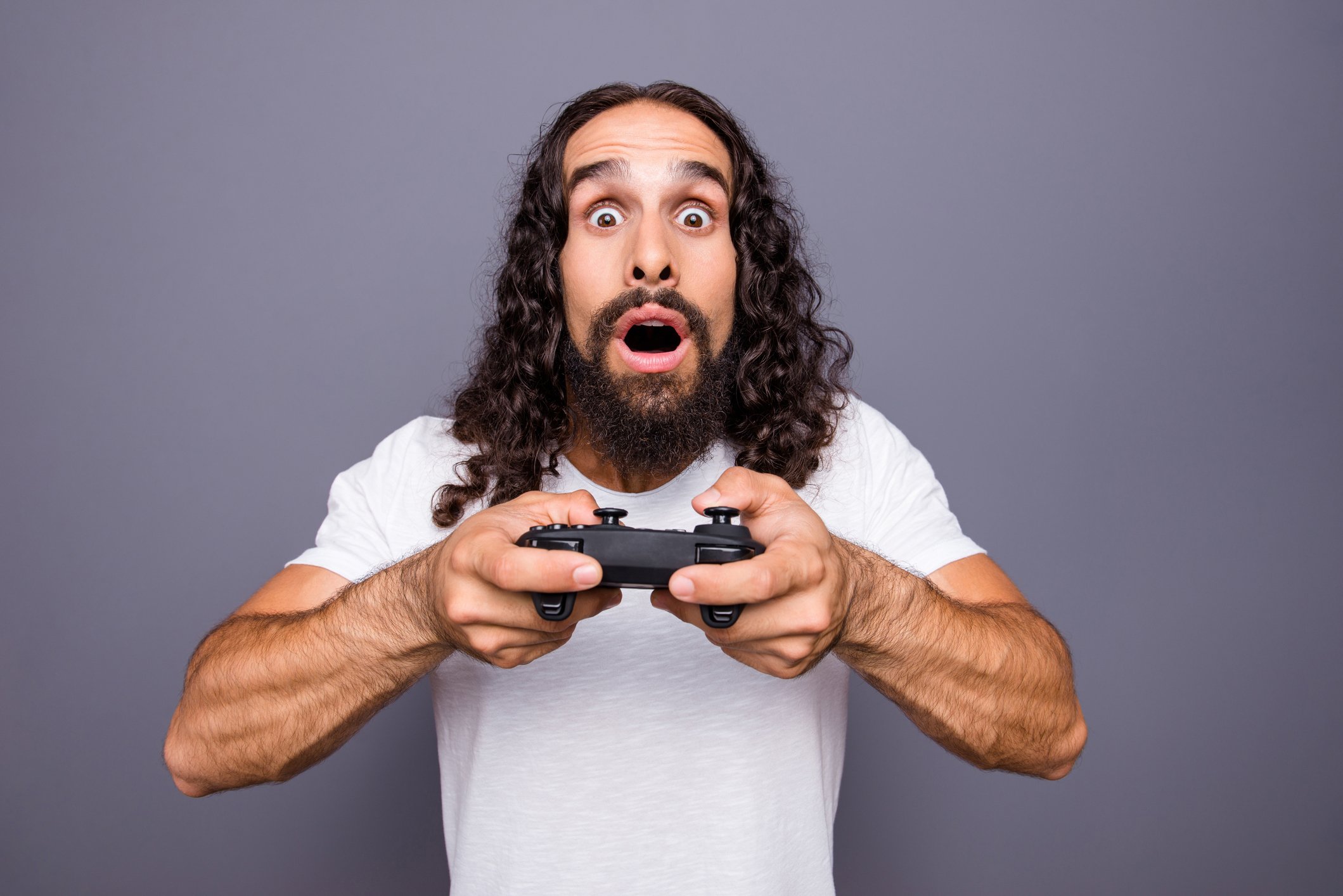 A man with long hair holds a gaming controller and is surprised facing into the camera. 