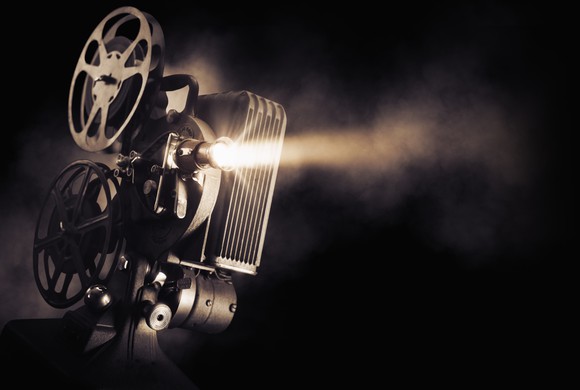 An old-school movie projector shoots light as reels of film circle above the projector. 