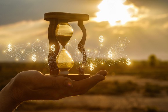 An open hand holding an hourglass with seven glowing dollar signs hovering around it. 