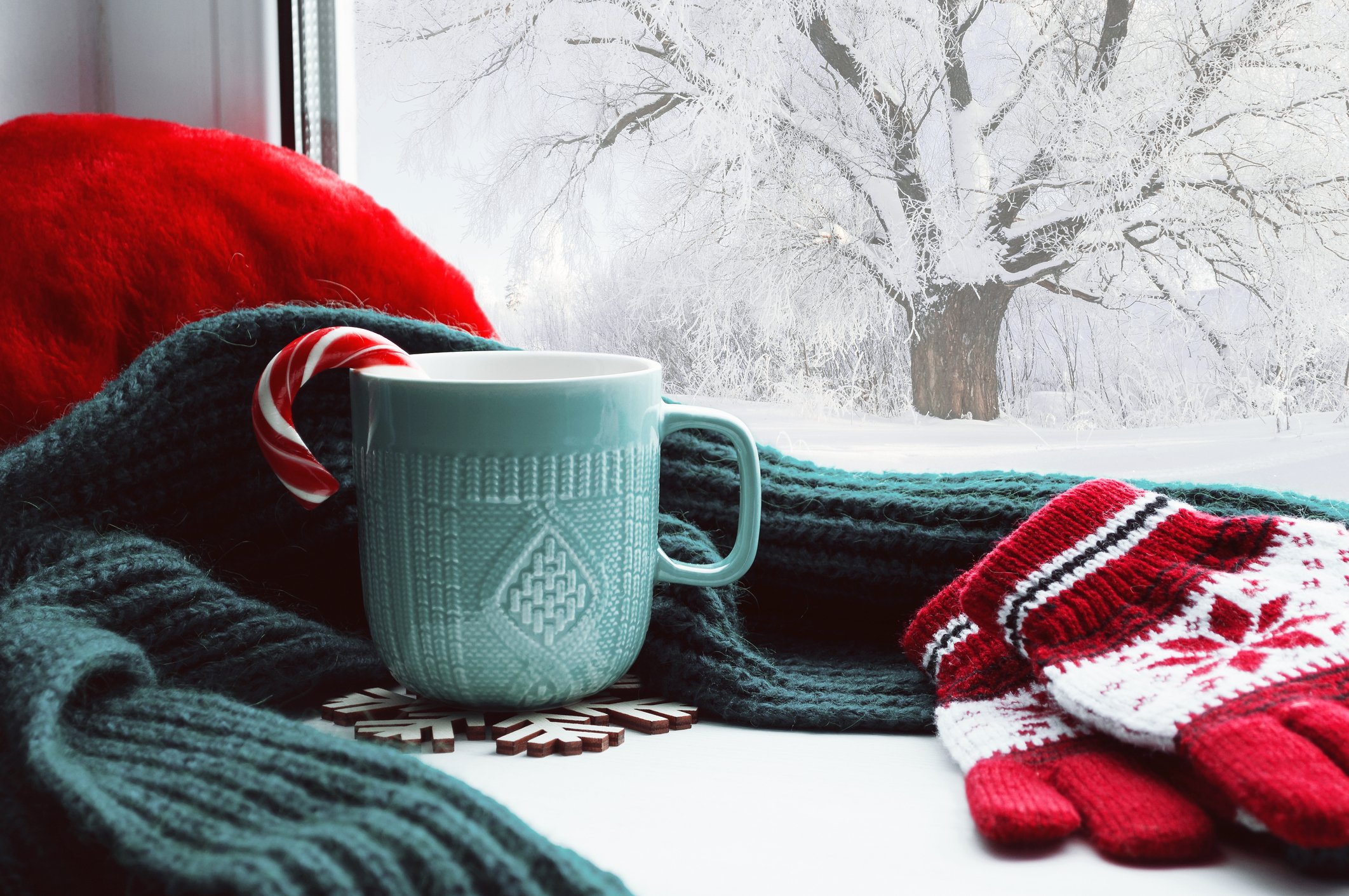 Winter scene through a window, with snow outside, and gloves, scarf, and mug of cocoa on the windowsill