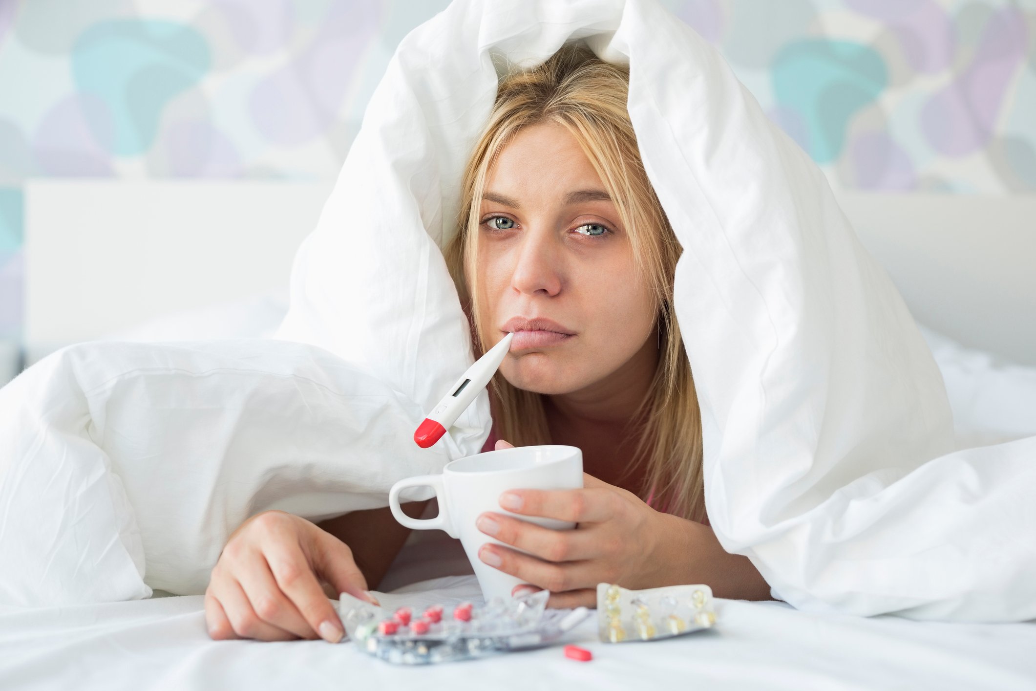 A sick young woman under the blankets with a thermometer in her mouth, a cup in her left hand, and over-the-counter medicine packets flanking her. 