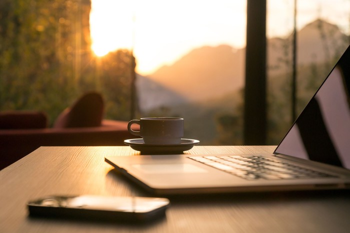A laptop, phone, and cup of coffee sit on a desk next to a window revealing a view of the sun and mountains.