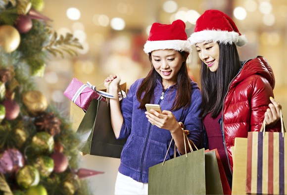 Two women wearing Santa hats, carrying shopping bags, and looking at something on a smartphone