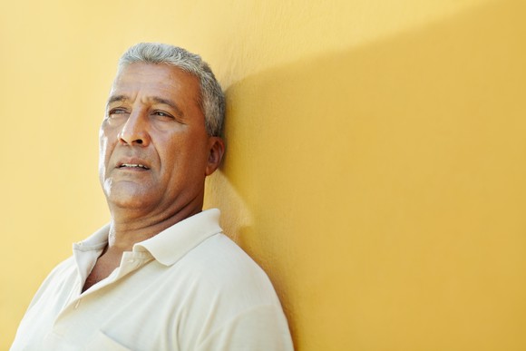 Older man with serious expression leaning against yellow wall