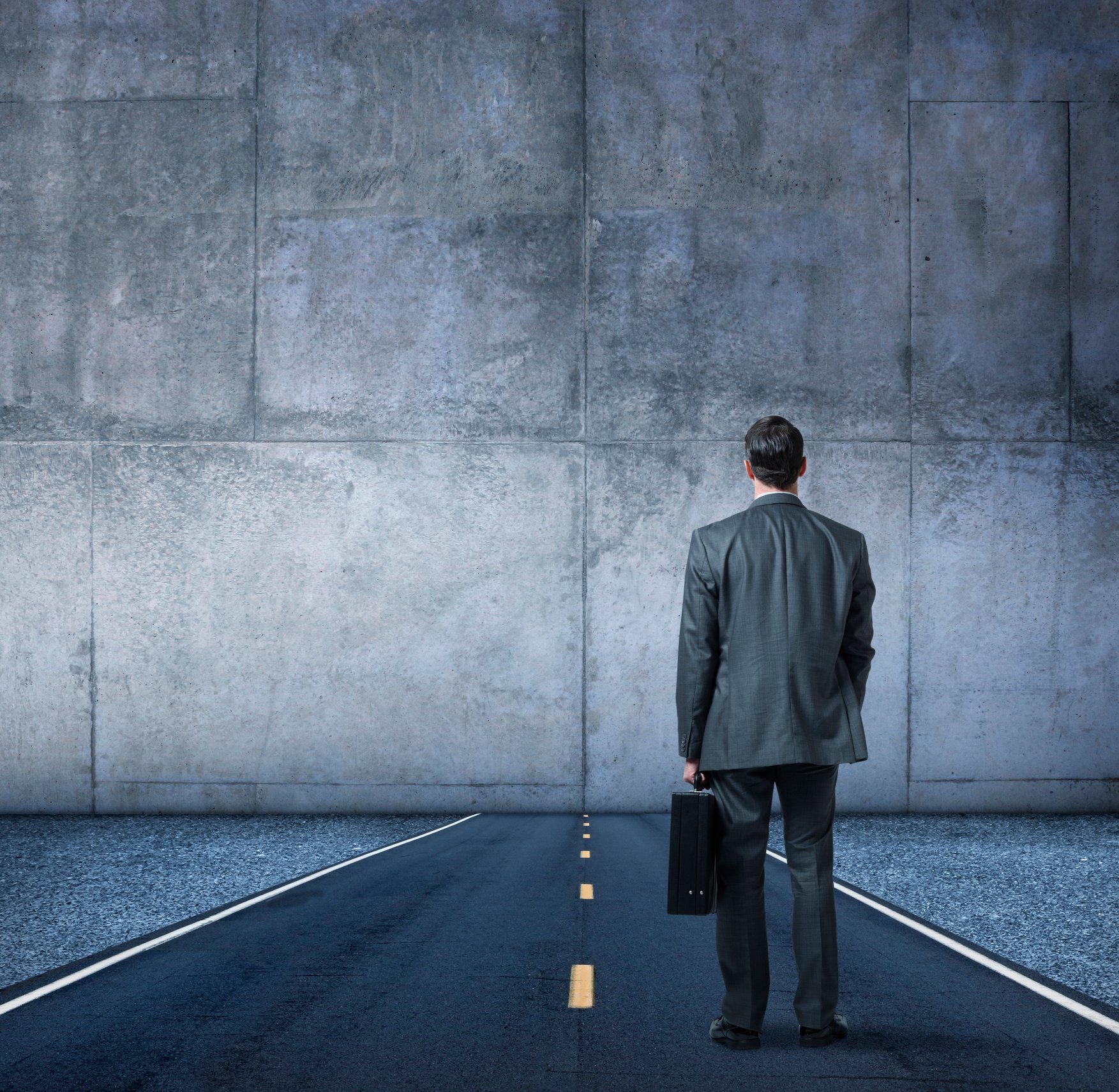 A man in a suit holding briefcase looking at a road blocked by a wall