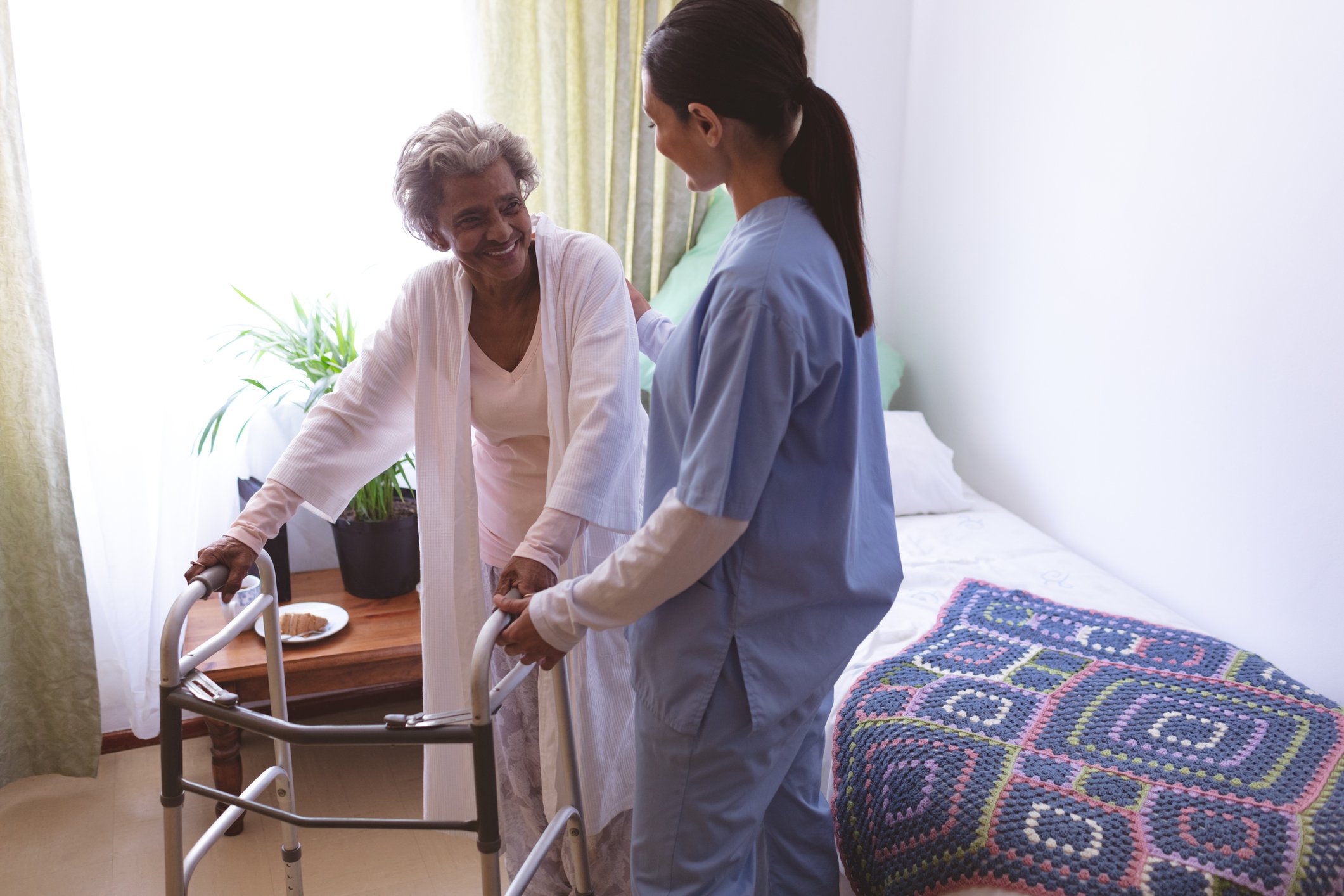 A woman in scrubs standing next to a bed, helping a woman with a walker.