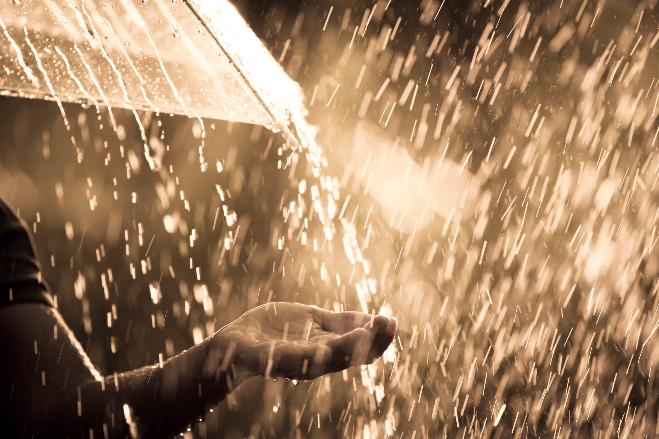 A person holds an umbrella in a heavy rainstorm, reaching the other hand out to catch some water.