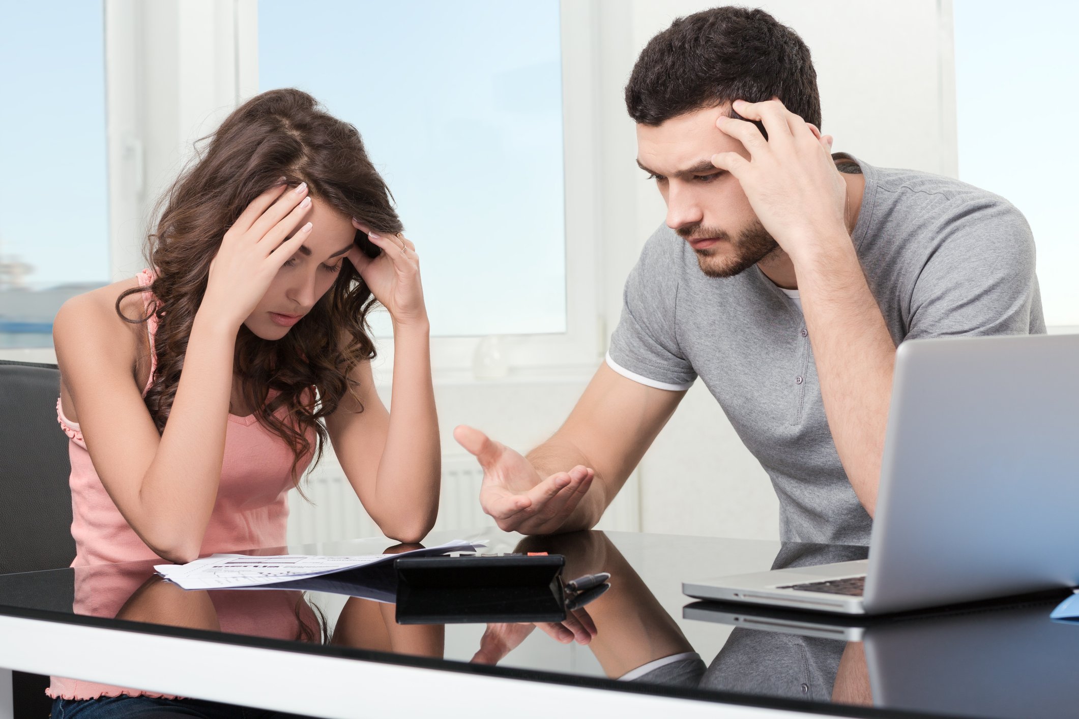 Young man at woman holding their heads; a laptop and documents sit on the table they're at