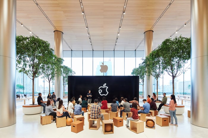 Large hall with trees and pillars, a big screen with Apple's logo on it, and about two dozen people watching a presentation.