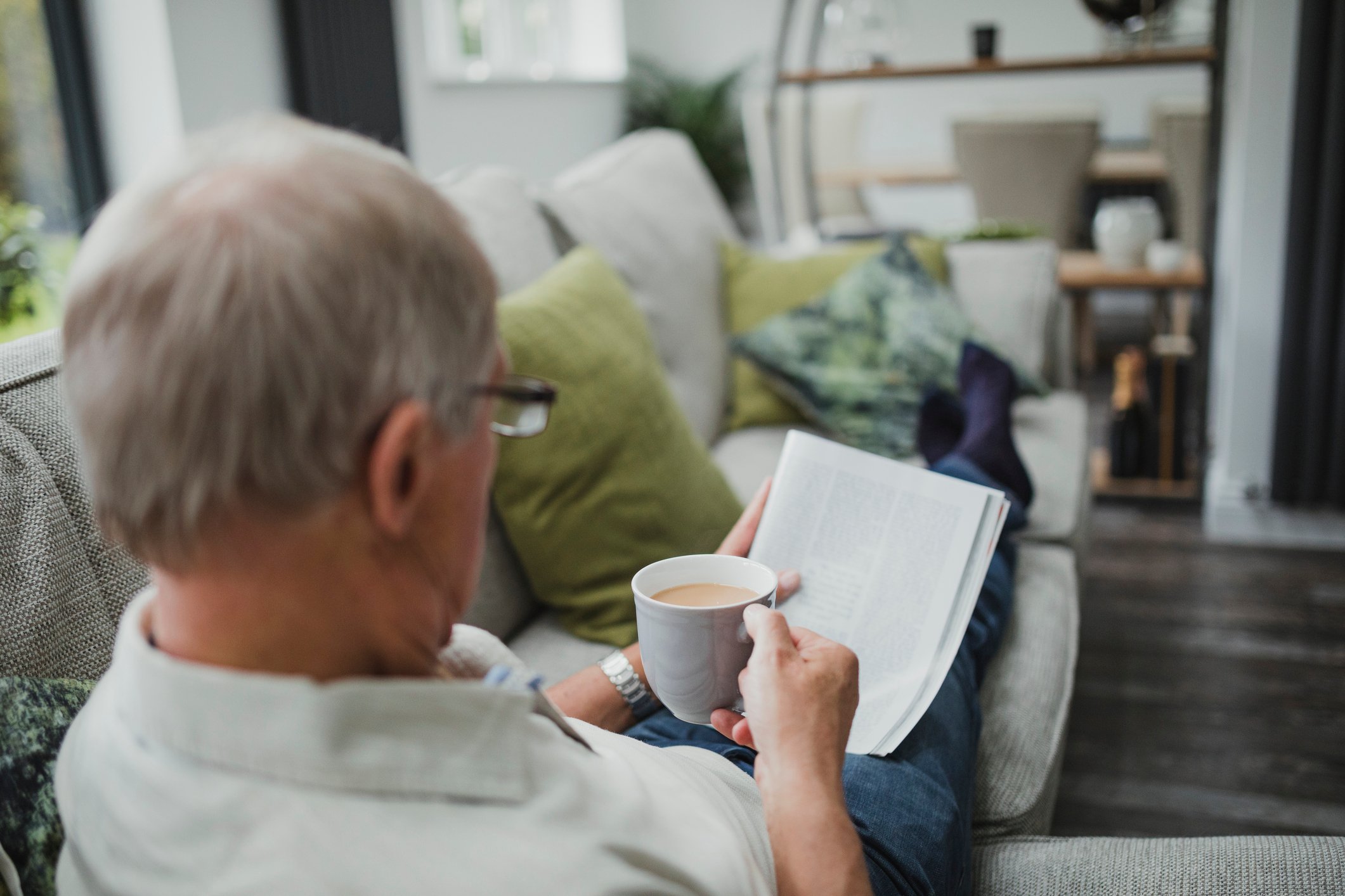 Gray-haired man reading a magazine while holding a mug and reclining on a couch.