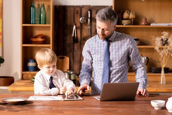 A man and a boy wearing ties look at a laptop, a tablet, and a printout on a table