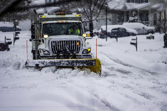 A work truck with yellow snow plow attachment clears a residential street after a winter storm.