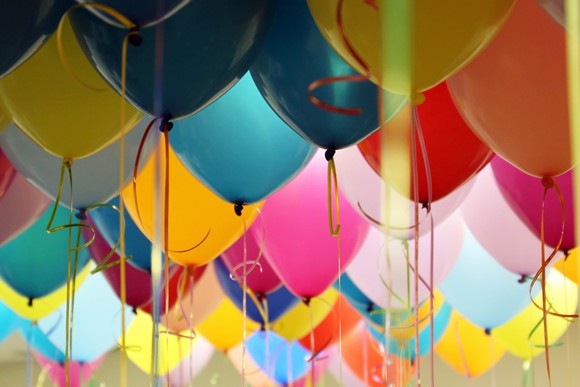 Helium-filled balloons in pink, yellow, and blue float together in a room.