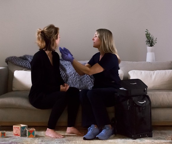 A woman in scrubs and medical gloves sitting on a couch checking a woman sitting next to her.