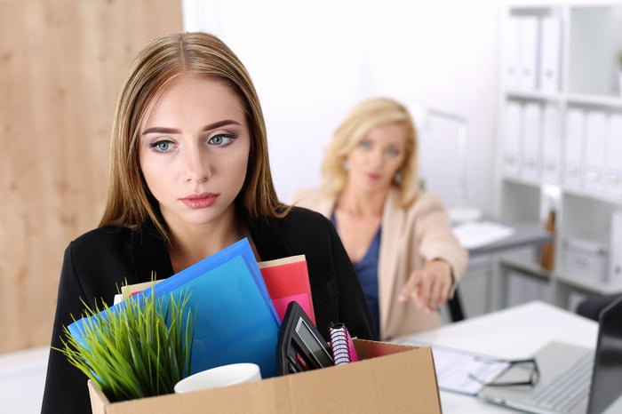 A professionally dressed woman sadly holds a box of belongings as another woman at a desk points at her to leave.