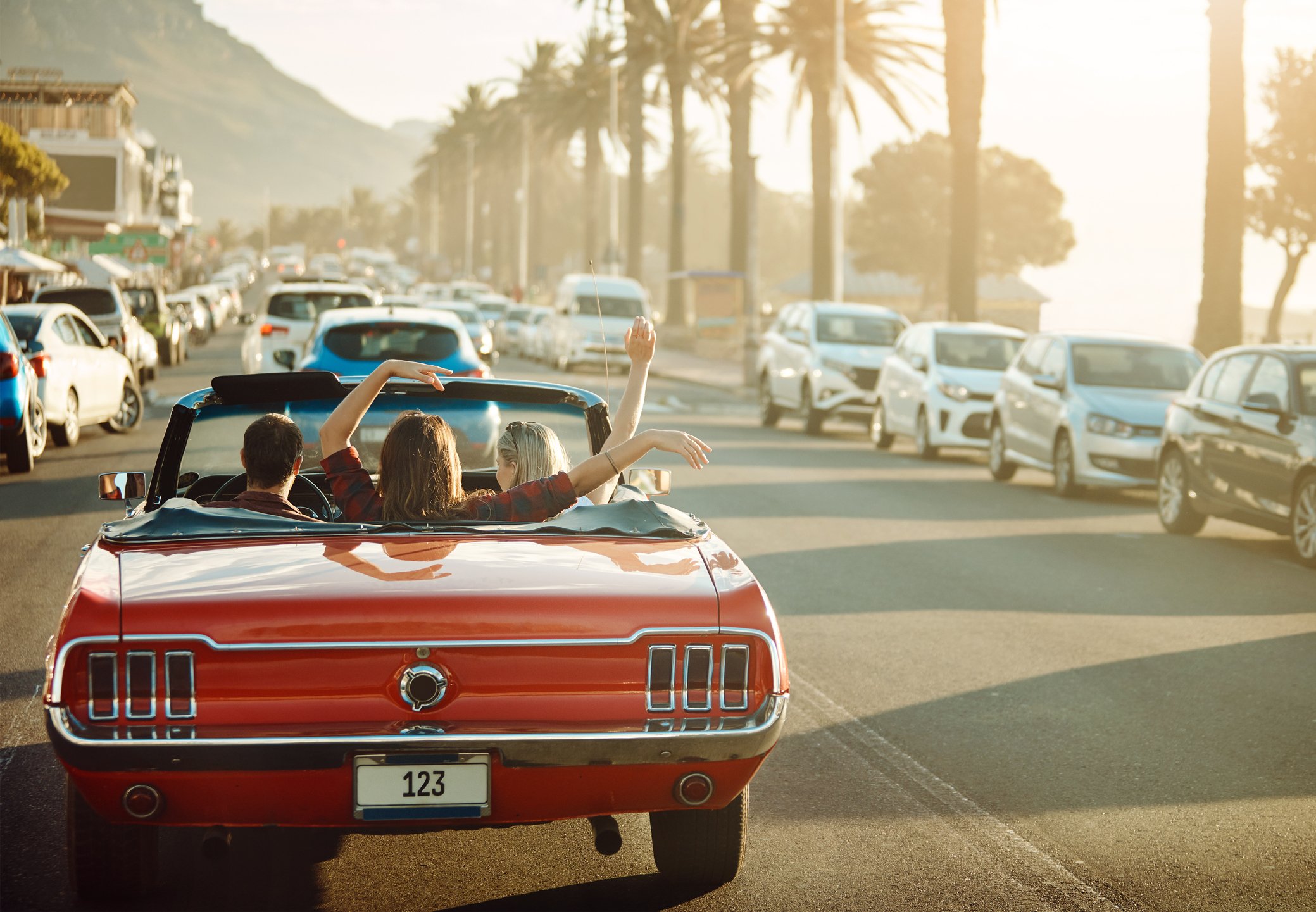 A red convertible on a road lined with palm trees, containing people waving their arms happily