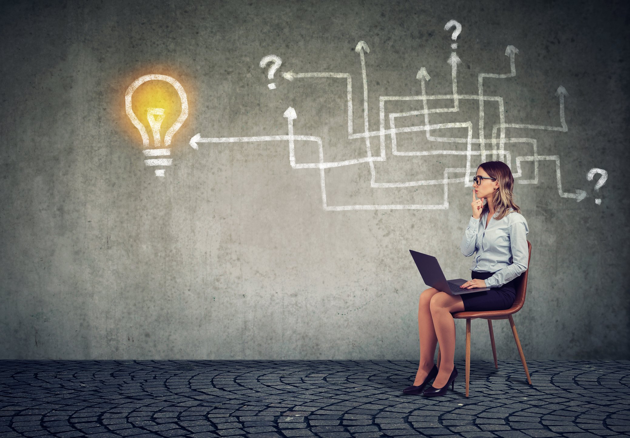 In front of a maze of arrows and question marks, a businesswoman looks at a light bulb on a wall.