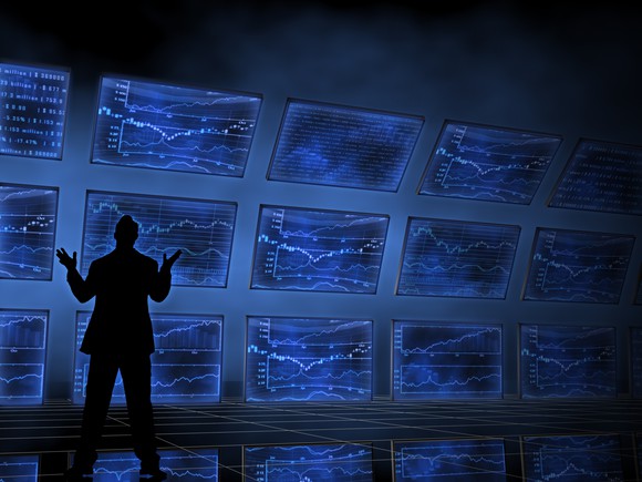 A silhouetted person in front of multiple blue-lit screens showing various charts and graphs