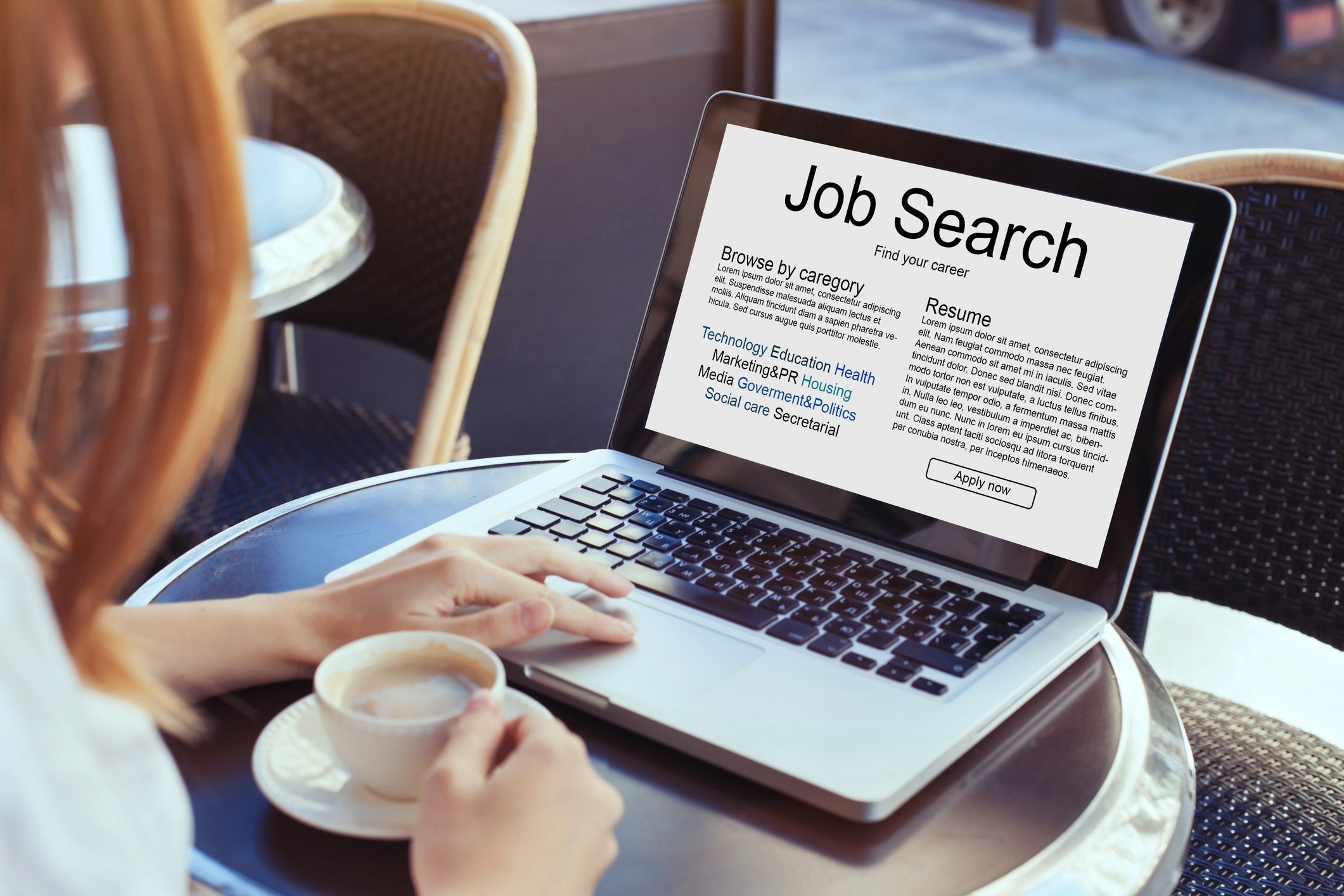 A woman sits at a small table in a restaurant drinking coffee in front of a laptop computer that says "Job Search"