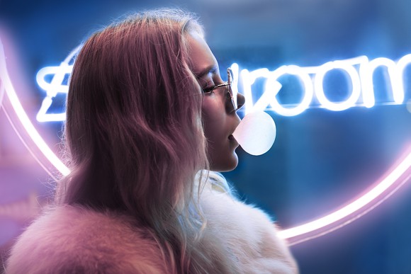 A young woman blows bubble gum in front of a neon sign.