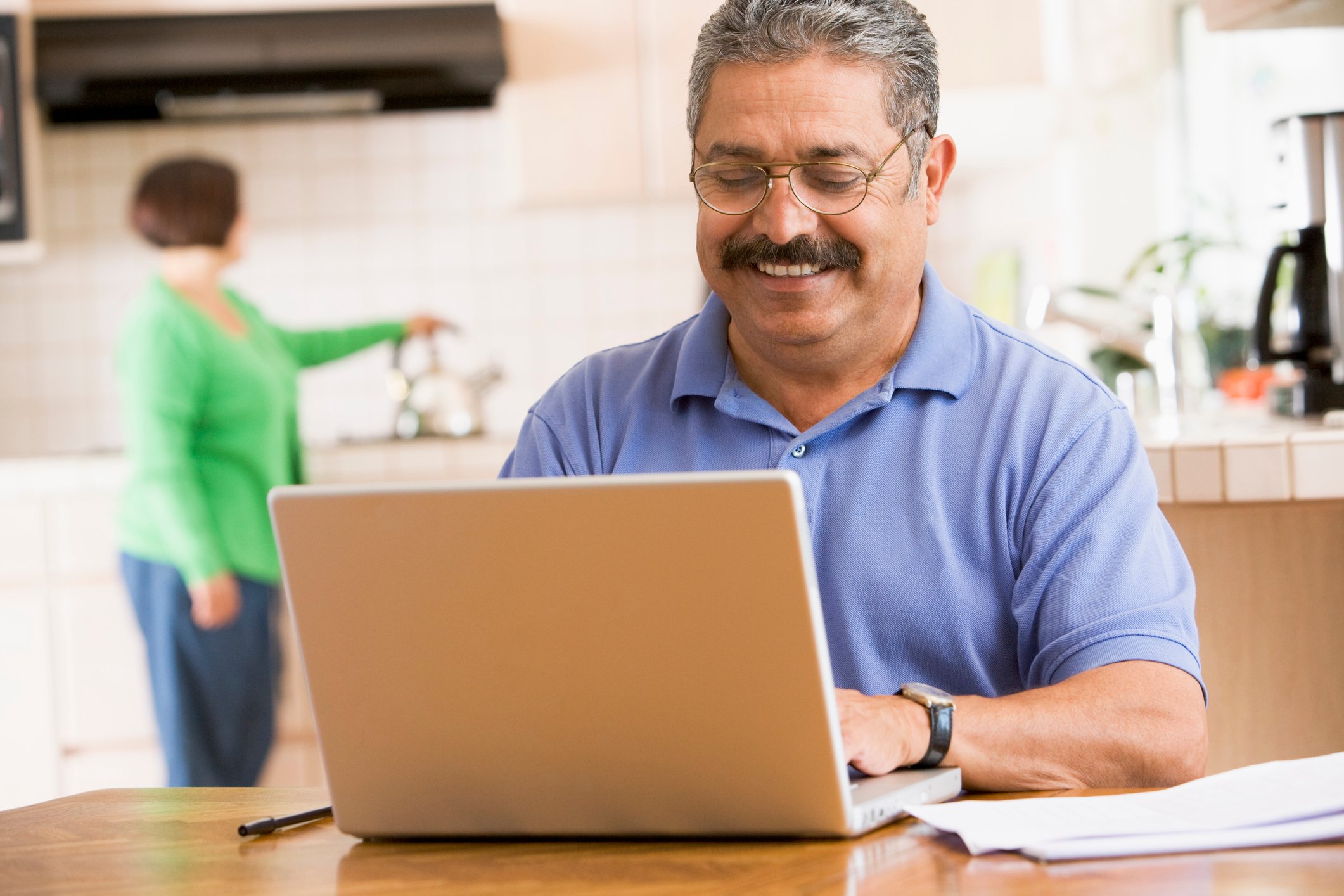 A smiling man on a laptop at the kitchen table, while a woman puts a kettle on the stove in the background.