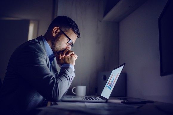 A man sitting in front of a laptop holding his hands to his mouth and looking pensive.