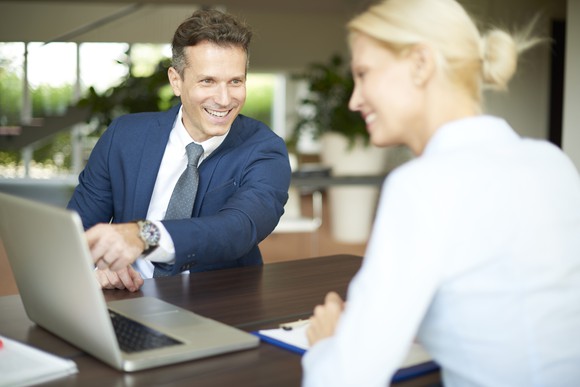 A smiling businessman points to his laptop screen and a smiling young woman follows along, pen and notebook ready to go.