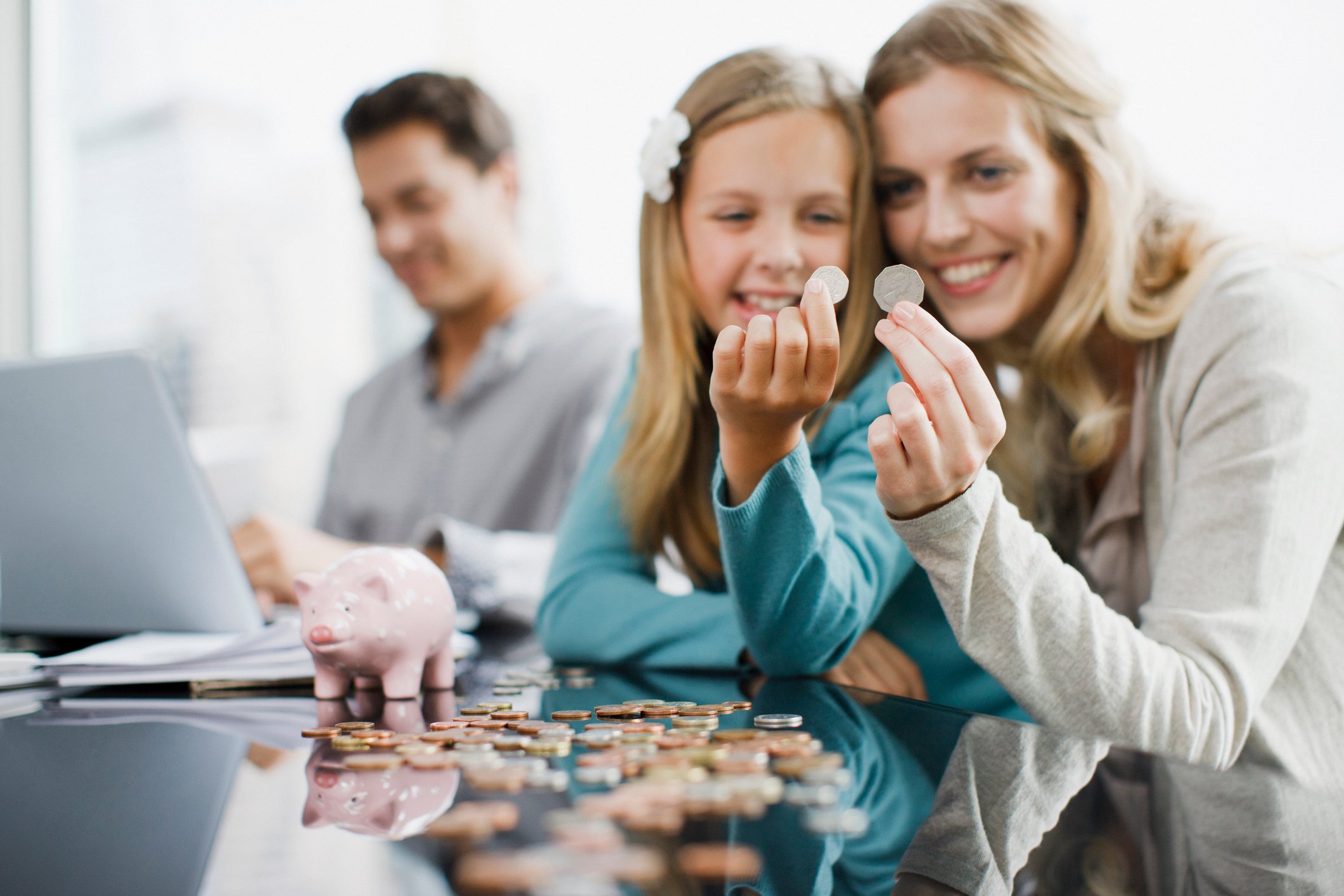 A young mother smiles and holds up coins with her daughter next to a piggy bank at a table while Dad operates his laptop behind them out of focus. 