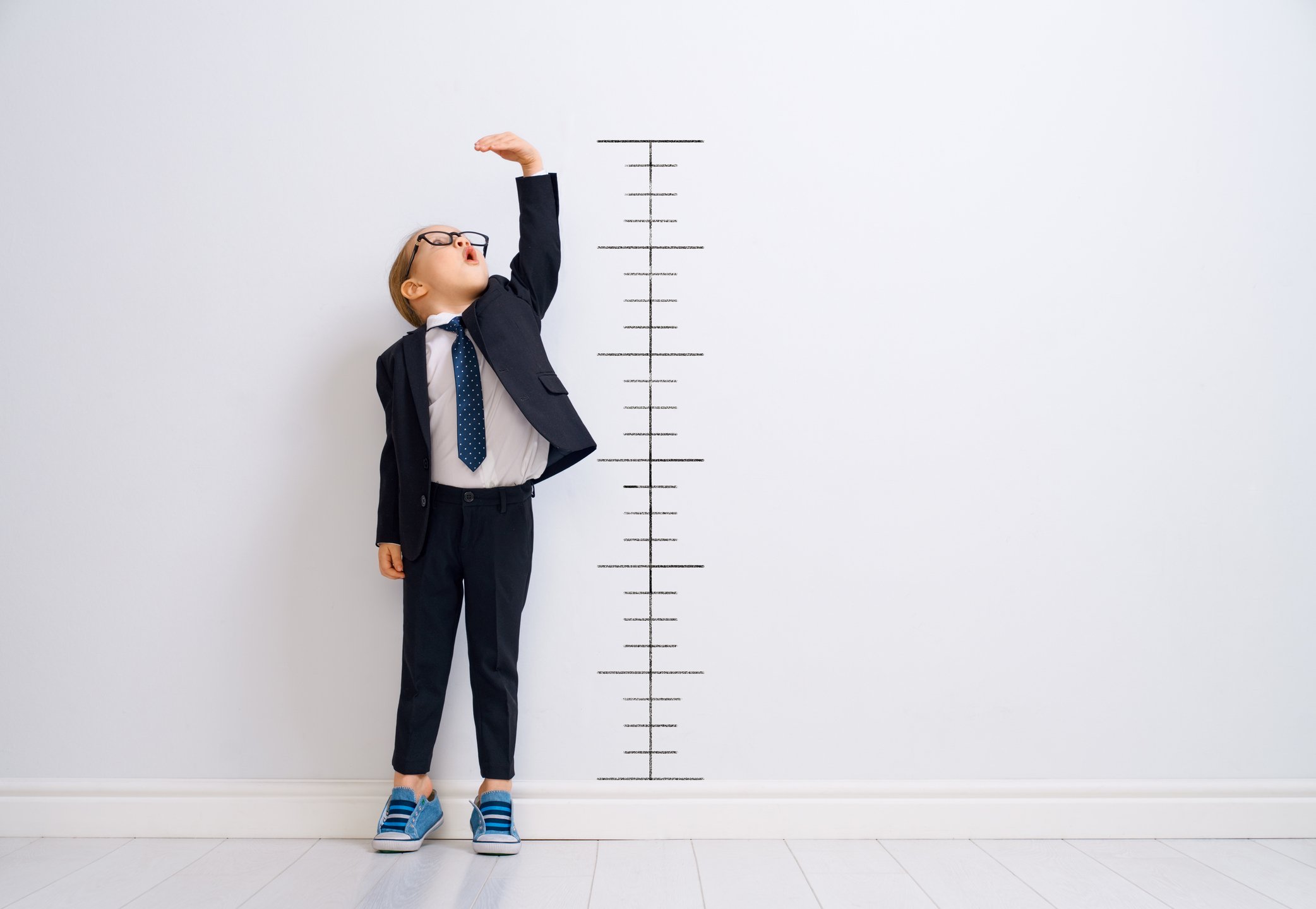 A young boy in a business suit standing in front of a wall on which there are hash marks indicating height