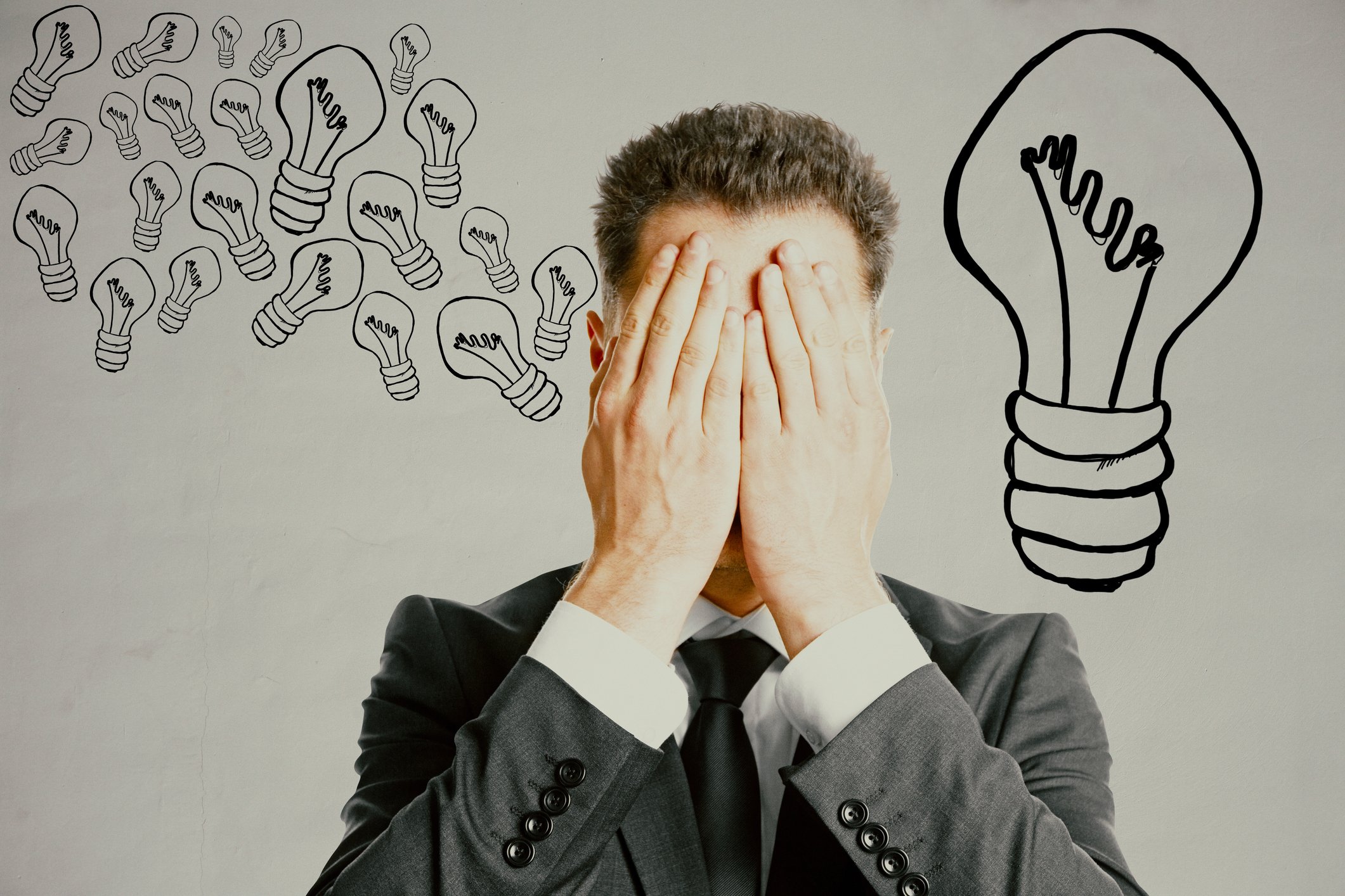 A man covers his eyes with the palms of his hands while standing in front of drawings of light bulbs on a wall.