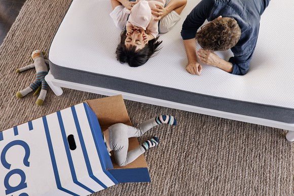 A man, woman, and baby on a mattress next to a toddler crawling into an empty cardboard box from Casper.