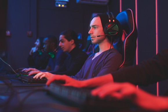 A group of people sit together at a long table playing PC games