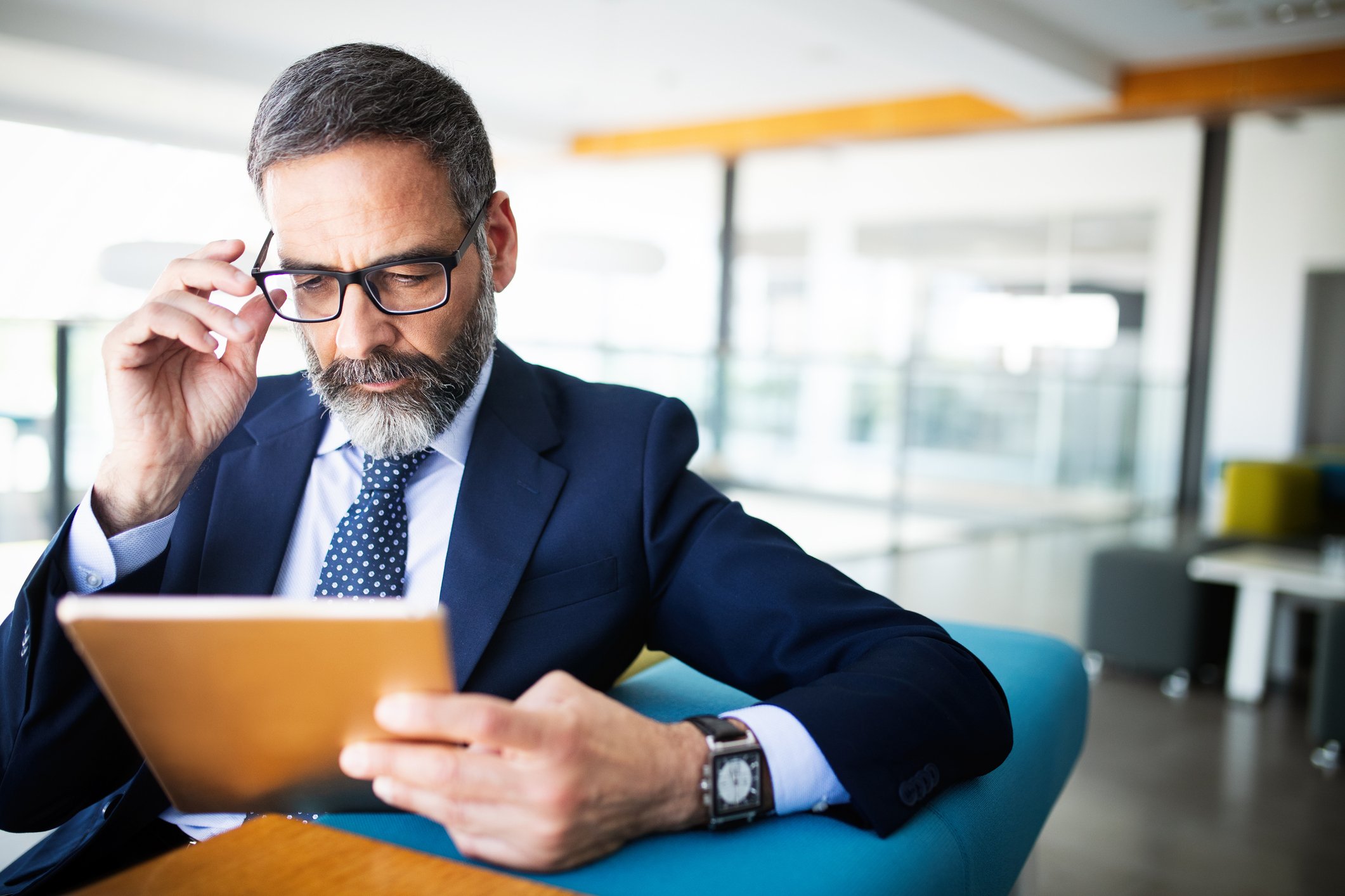 Adjusting his glasses, a man in a suit reads his tablet.
