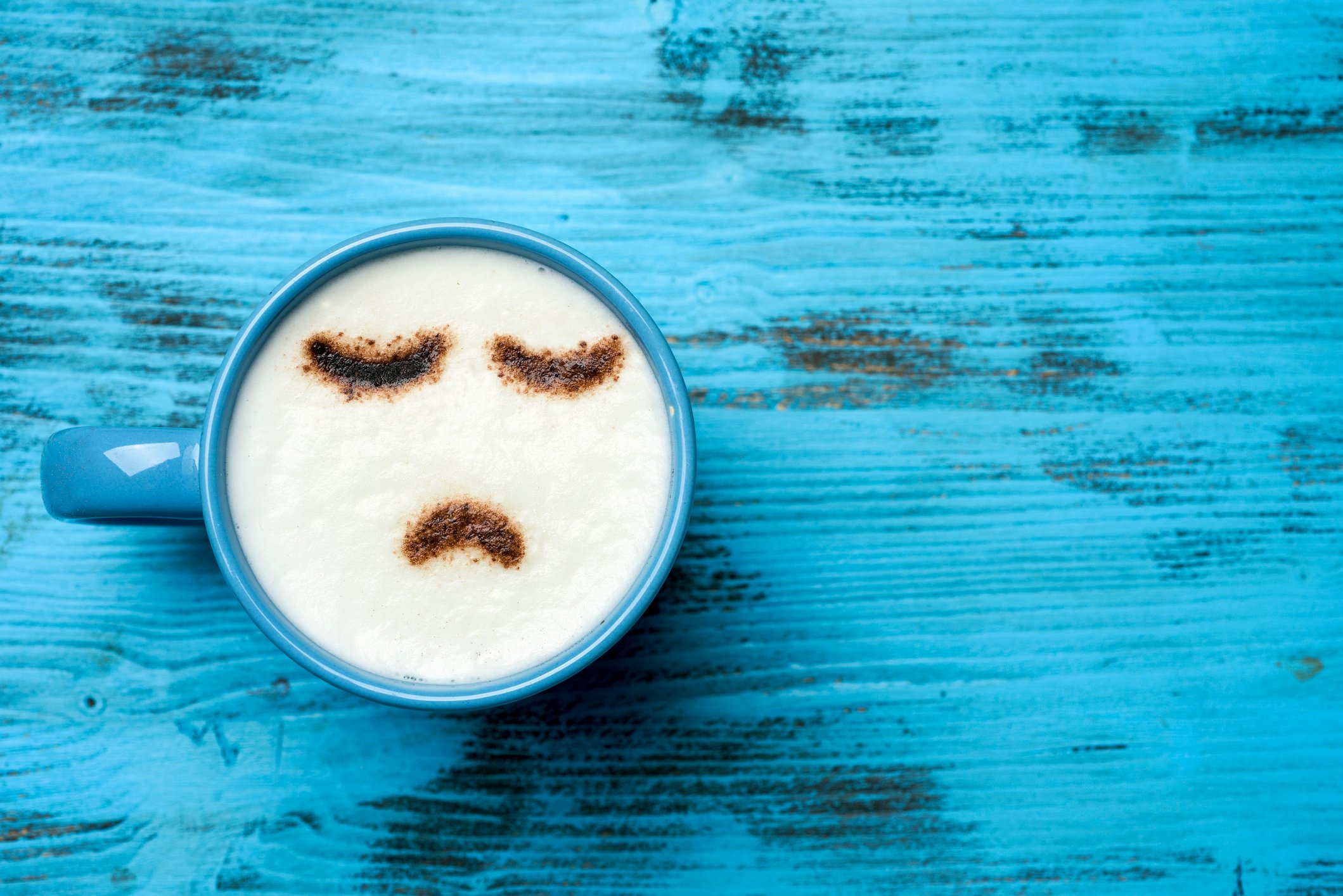 high-angle shot of a blue cup of cappuccino with a sad face drawn with cocoa powder on its milk foam