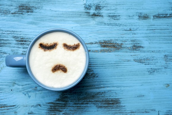 high-angle shot of a blue cup of cappuccino with a sad face drawn with cocoa powder on its milk foam