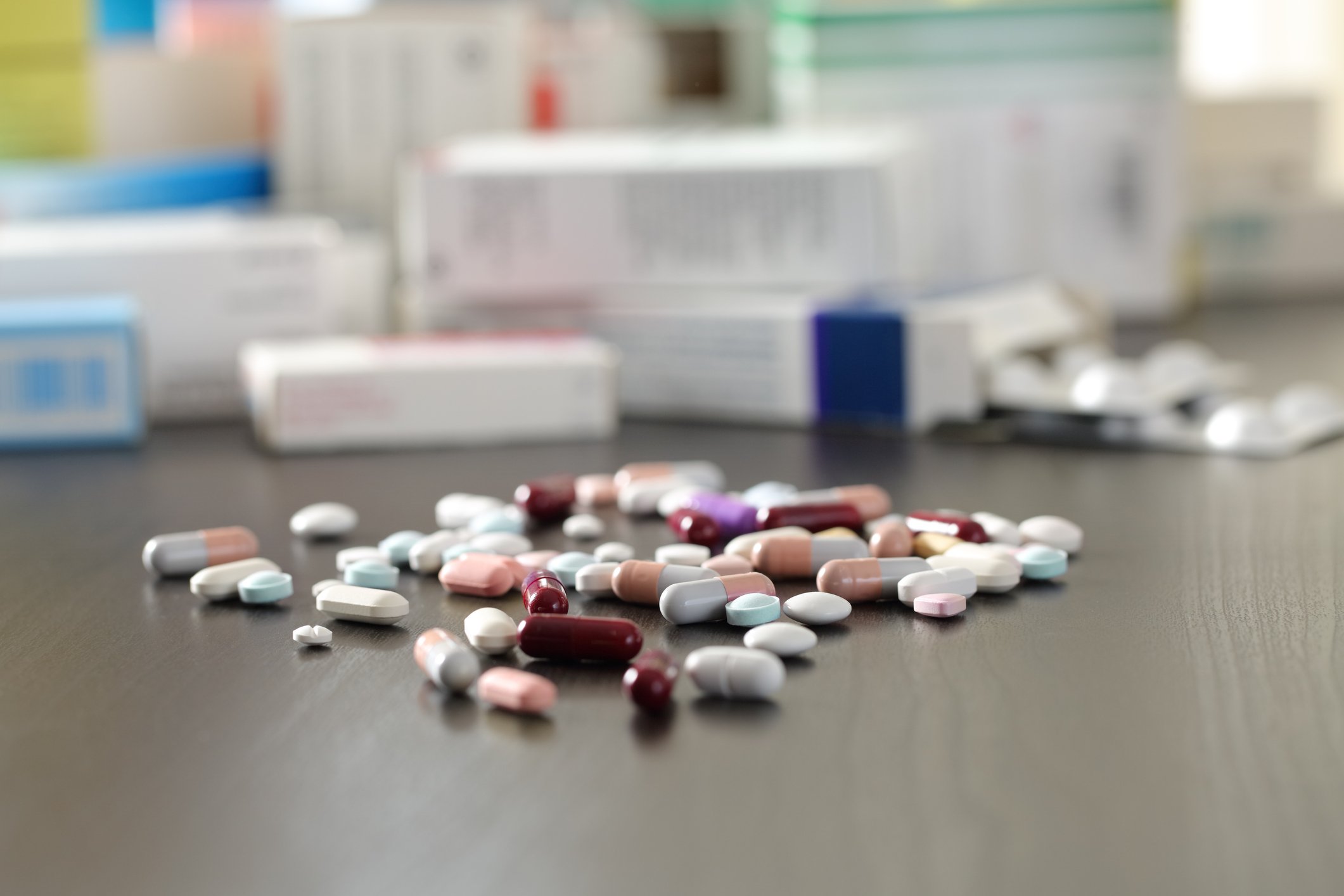 A variety of pills are spilled onto a table with empty medication boxes in the background.