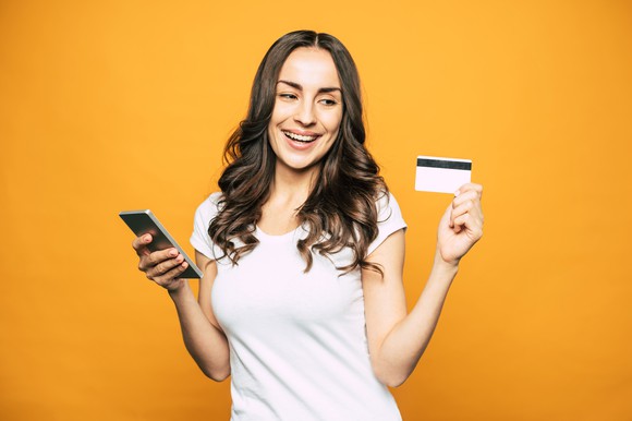 A young woman smiles and holds a credit card in one hand and her cell phone in the other. 
