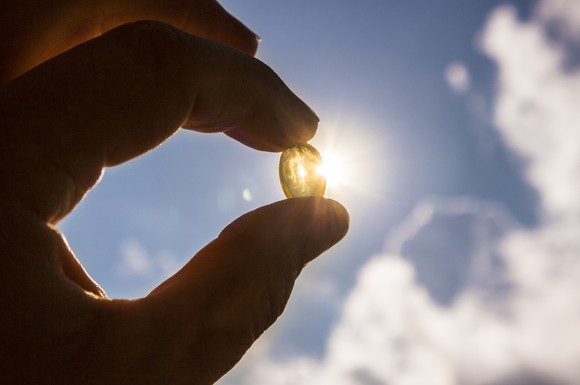 A person's hand holds a fish-oil based pill up to a blue sky with sun reflecting off the pill.