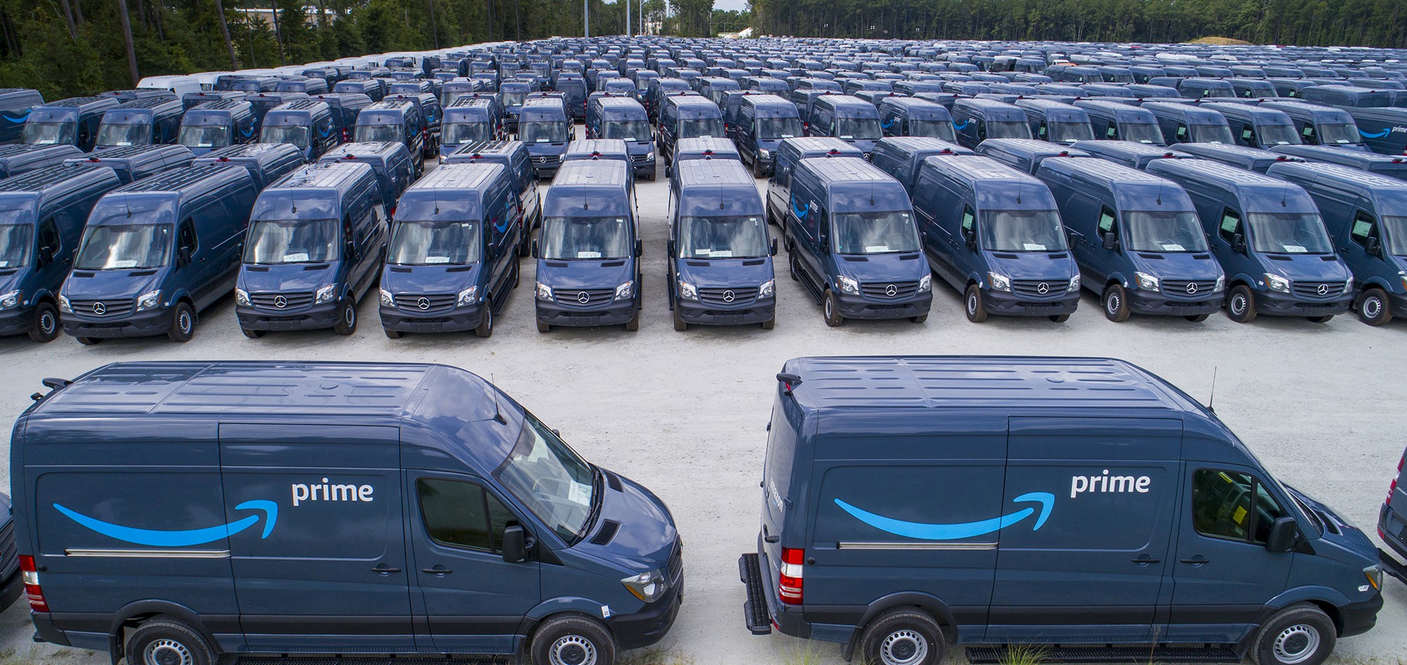 Several dozen dark blue Amazon Prime delivery trucks lined up in a large parking lot.