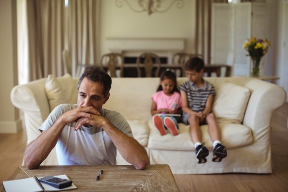 Man with serious expression rests his elbows on a table with a pad of paper and calculator, while a boy and girl sit on a couch in the background