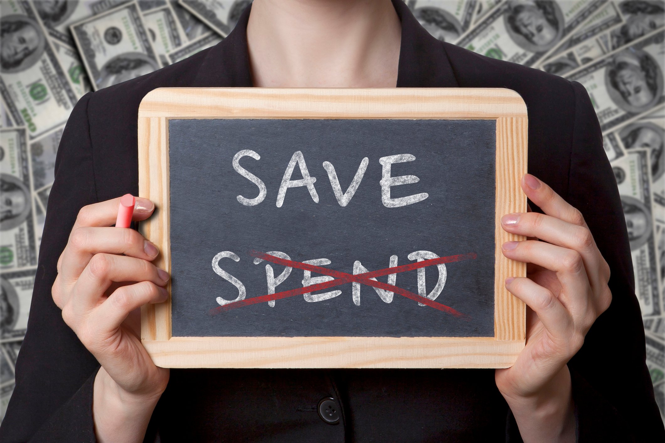 Someone holding up a small blackboard, on which is printed the words "save" and "spend," with the word "spend" crossed out.