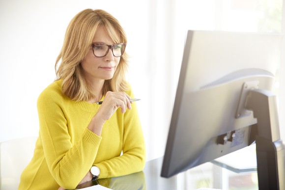 A middle-aged woman in yellow sweater looks at a large desktop monitor with a pen in her hand.