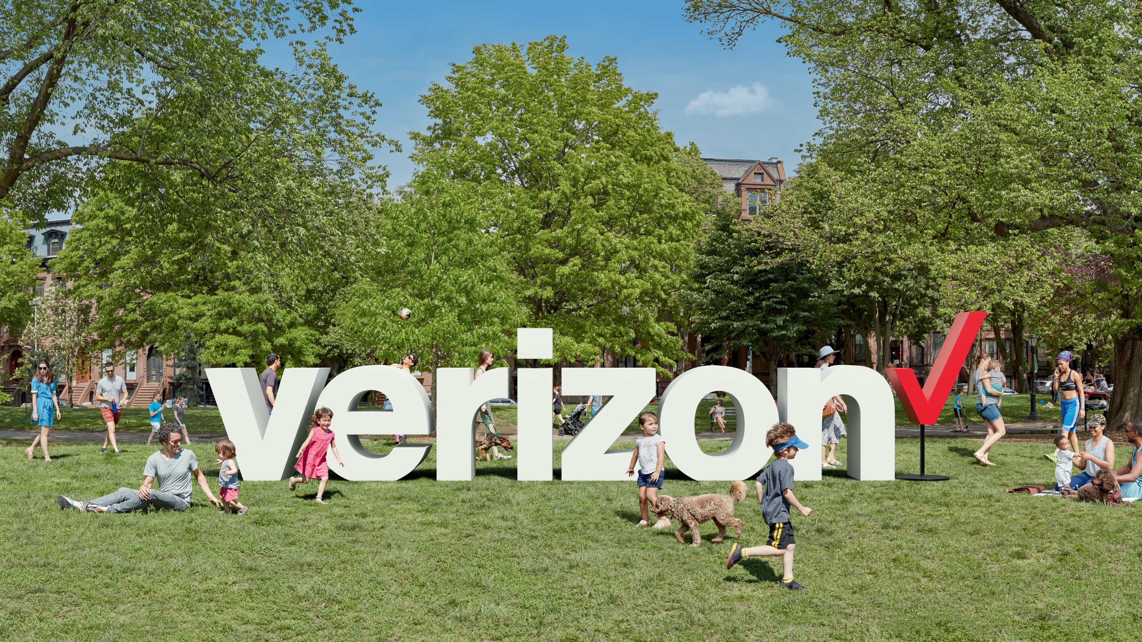 A 3D model of Verizon's logo superimposed over a picture of a park filled with people playing and relaxing